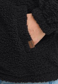 Svart, strukturert fleecejakke med en stor håndlomme. Har en brun lærmerkelapp med "oymto"-logo nær lommeleddet.