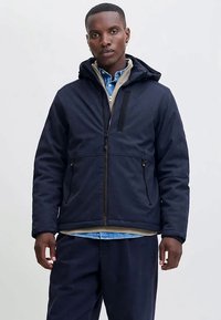 Tamsiai mėlynas bliuzonas su gobtuvu, su užtrauktuku priekyje, tekstūruota medžiaga, šoninės kišenės ir šviesiai pilki akcentai. Dėvimas ant mėlynos langelinės marškinės.