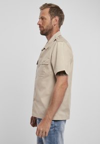 Camicetta beige a maniche corte realizzata in tessuto con una texture liscia, dotata di due tasche sul petto e una chiusura frontale con bottoni, rifinita con colletto.