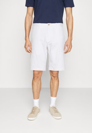 Mann trägt weiße kniehohe Shorts, dunkelblaues T-Shirt, weiße Socken und beige Turnschuhe und steht vor einem einfarbigen Hintergrund.
