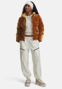 Veste matelassée en velours brun avec doublure jaune, associée à un pantalon élastique blanc avec poches zippées noires et baskets beiges.
