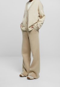 Cardigan beige con scollo a V, maniche a costine e grandi bottoni, abbinato a pantaloni beige a gamba larga. Calzature con suola dal design testurizzato.