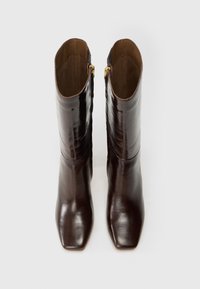 Par de botas de cuero pulido marrón oscuro hasta la mitad de la pantorrilla con cremalleras laterales y punteras cuadradas, vistas desde arriba sobre un fondo blanco.
