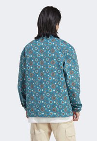 Blå mönstrad jacka med blommönster i vitt och brunt. Lättviktig tyg, långa ärmar och avslappnad passform, som slutar vid höfterna.