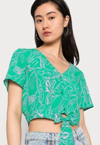 Top verde curto com mangas curtas, apresentando um design com laço na frente e padrões florais abstratos em rosa. Decote em V e detalhe dos botões visíveis.