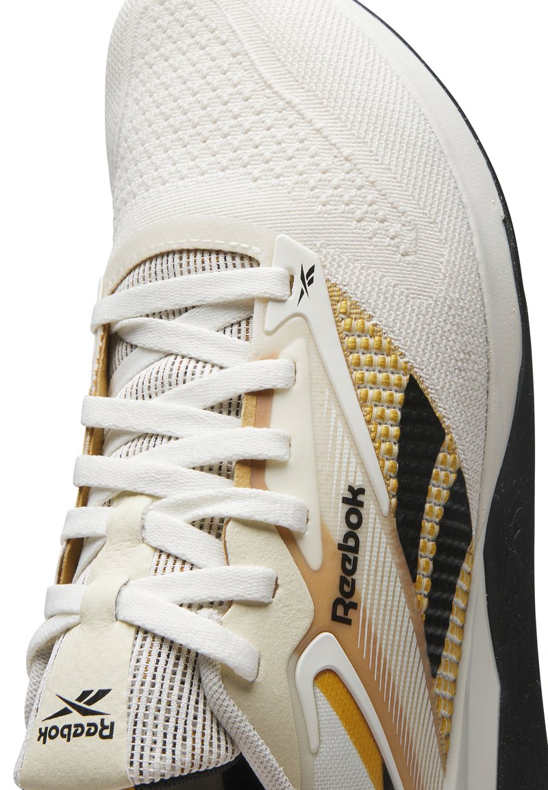 Zapato deportivo blanco con parte superior de malla texturada, acentos en dorado y negro, sistema de cordones destacado y logotipo de Reebok visible.