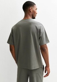 Grå bomullströja med avslappnad passform, korta ärmar och en enkel design. Bakvy visar en rak fåll och minimala sömnadsdetaljer.