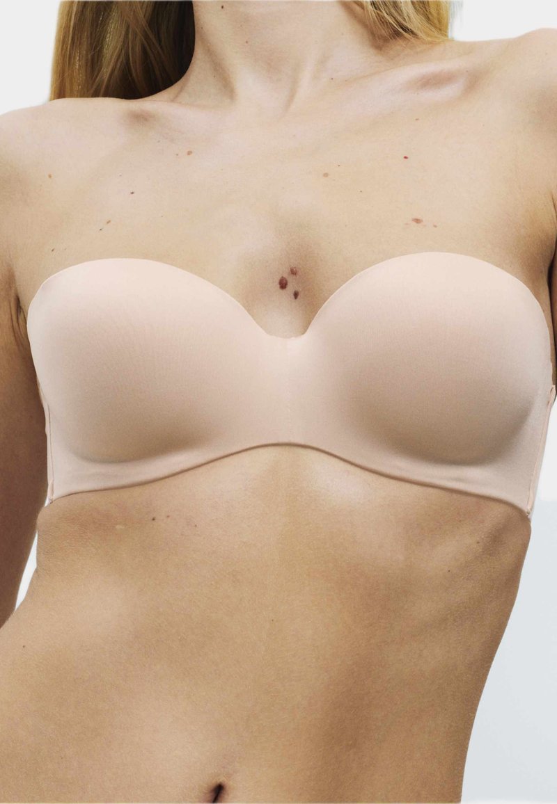 Woman wearing a nude strapless bra, showing upper torso and neck with visible small skin moles and light skin tone.