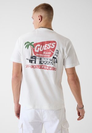 Hombre con camiseta blanca con logotipo estilo vintage en rojo, negro y verde de "Guess Jeans California", con gráficos de palmera, edificio y mujer en la parte trasera.