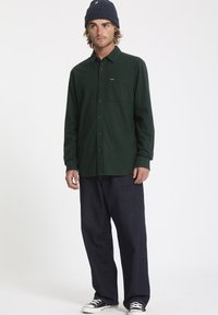 Chemise boutonnée vert foncé avec une poche poitrine, associée à un jean bleu foncé ample et des baskets noires. Le modèle porte un bonnet bleu marine.