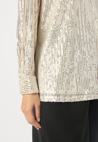 Maglione lungo beige chiaro a maniche lunghe decorato con paillettes argentee disposte in linee verticali, con polsini abbottonati e una texture liscia.