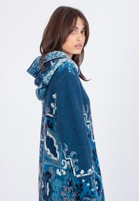 Blauwe gebreide hoodie met een textuurpatroon en geometrische en bloemenmotieven. De capuchon heeft een contrasterend patroon langs de rand.