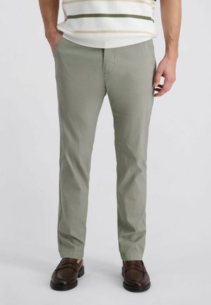 Mann trägt hellgraue, schmal geschnittene Hose, braune Leder-Loafer und ein weißes Hemd mit grünen Streifen, steht mit den Händen in den Taschen.