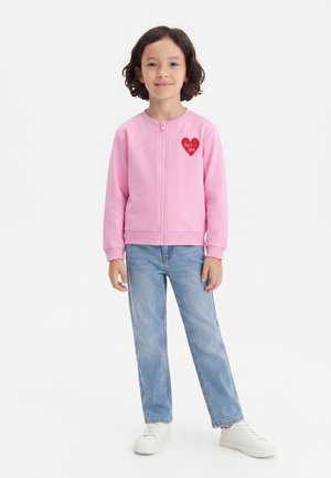 Barn med mørkt krøllet hår iført en pink jakke med lynlås og et rødt hjerte-patch med teksten "Self Love,", blå jeans og hvide sneakers, står.
