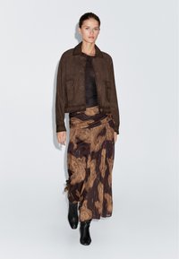 Frau trägt eine dunkelbraune Wildlederjacke, ein schwarzes Oberteil, einen gemusterten braunen Midirock mit goldenen Akzenten und schwarze Stiefel mit Absatz, geht nach vorn.