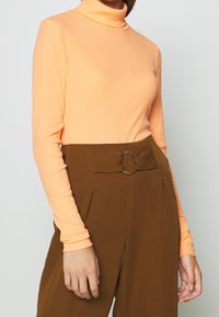 Langärmliger Rollkragenpullover aus pfirsich-orangefarbener Rippware, kombiniert mit hochgeschnittenen braunen Hosen, die mit einem runden Schnallen-Detail versehen sind.