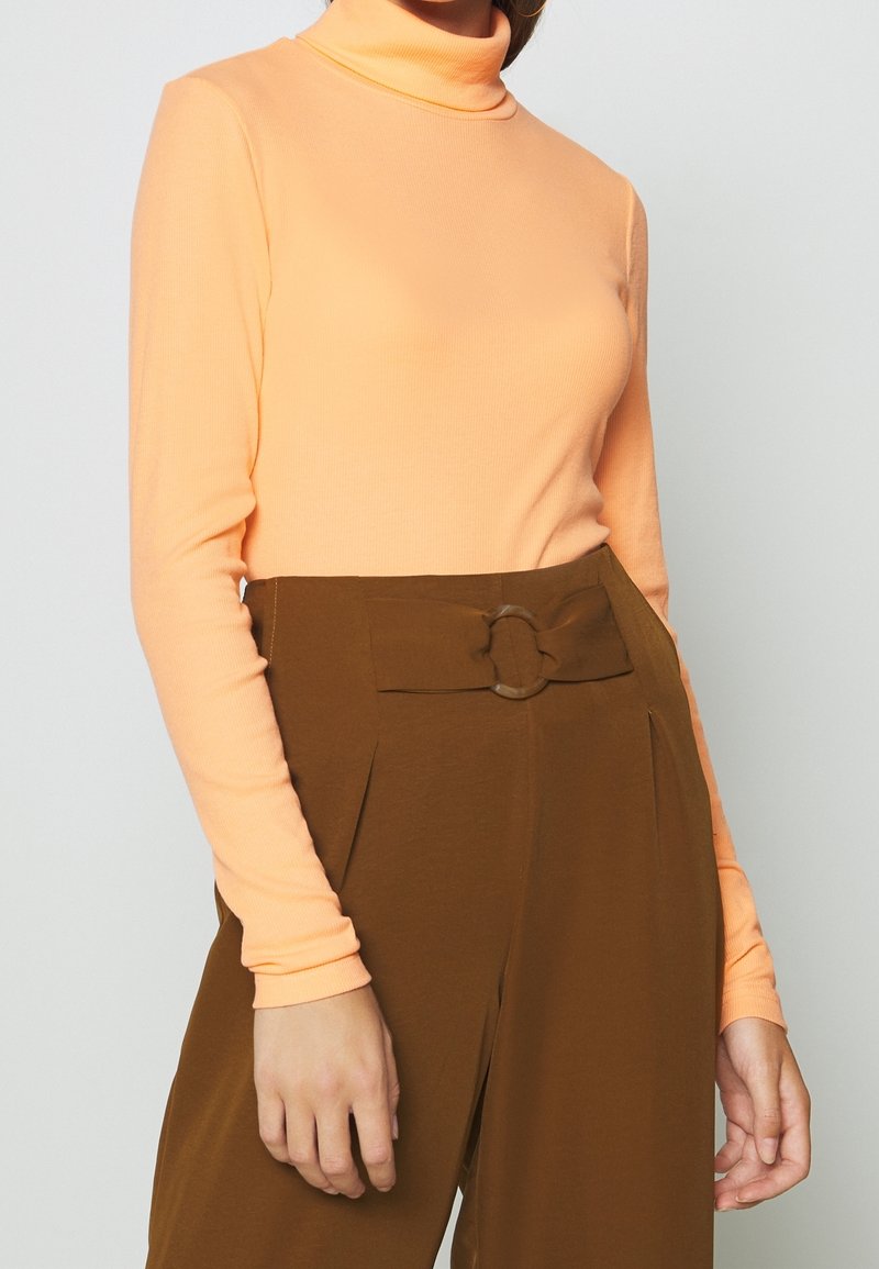 Langärmliger Rollkragenpullover aus pfirsich-orangefarbener Rippware, kombiniert mit hochgeschnittenen braunen Hosen, die mit einem runden Schnallen-Detail versehen sind.