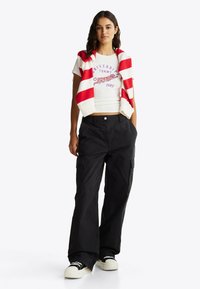 Top corto blanco con un gráfico morado y el texto "Universidad Tommy 1985", combinado con pantalones cargo de corte holgado en negro y un suéter a rayas rojas.