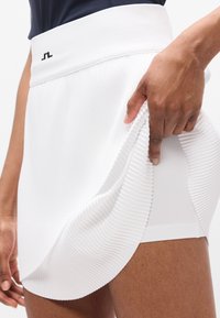 Λευκή πλισέ φούστα τένις με υφασμάτινη ζώνη με υφή. Το ύφασμα είναι λείο και ευλύγιστο, με κυρτό τελείωμα και διακριτική λεπτομέρεια λογότυπου.