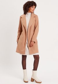 Kamel-farbener Mantel mit Reverskragen und langen Ärmeln, kombiniert mit einem hellen Strickkleid und cremefarbenen Plateaustiefeletten. Angenehme Oberfläche.