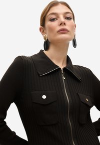 Vestido negro de canalé con cremallera frontal, gran cuello y dos bolsillos frontales, con detalles de botones en tono dorado y una silueta ajustada.