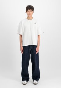 En ung mand står oprejst, iført en løs hvid struktureret T-shirt, mørke vide jeans og hvide sneakers foran en ensfarvet hvid baggrund.