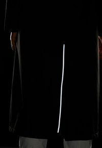 Manteau long noir avec une bande réfléchissante blanche droite le long de la couture arrière, doté de manches longues et d'une coupe ample.