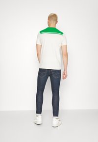 T-shirt de manga curta, na cor creme, com detalhes em verde nos ombros, combinada com jeans escuros de corte slim e sapatilhas brancas.