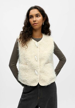 Crèmekleurige wolmix vest met een getextureerde, pluizige afwerking, voorzien van drie grote knopen en twee zijzakken. Draagt over een grijs langemouwen shirt.