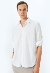Camisa de lino blanca de botones con mangas remangadas, que presenta un cuello clásico, tejido texturizado y un corte relajado.