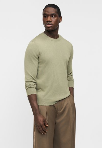 Homme portant un pull à manches longues vert clair et un pantalon marron ample, debout avec une main dans la poche, regardant sur le côté.