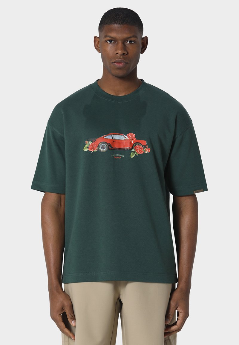 Homme portant un t-shirt oversize vert foncé avec un motif de voiture vintage rouge et de roses, associé à un pantalon beige, faisant face à l'avant sur un fond blanc.