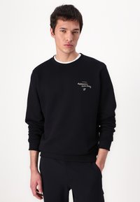 Black sweatshirt with crew neck and long sleeves. Features embroidered logo on chest. Soft texture, fitted cuffs and hem. Paired with navy pants.