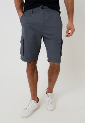 Mann trägt dunkelgraue Cargo-Shorts mit Klappentaschen und weiße Sneaker, steht vor einem einfarbigen hellen Hintergrund.