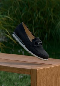 Μαύρο δερμάτινο μοκασίνι με διακοσμητική αγκράφα, λευκή σόλα και μπεζ εσωτερικό. Ομαλή υφή, στρογγυλεμένη μύτη και αντίθεση με την ξύλινη επιφάνεια.