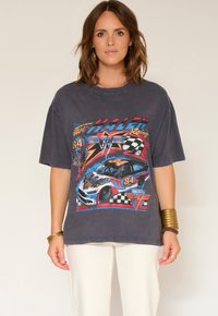 Femme portant un T-shirt oversize gris foncé avec un graphisme coloré d'une voiture de course vintage et le numéro "84", associé à un pantalon blanc et des bracelets en or.