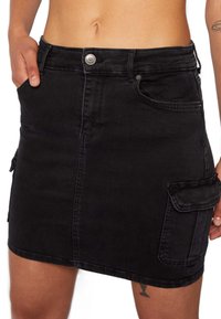 Jupe en denim noir avec poches plaquées, forme ajustée et fermeture à boutons à la taille. Le tissu présente une apparence lisse avec une légère texture.