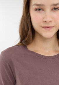 Brązowa, dopasowana koszulka z okrągłym dekoltem i gładką teksturą. Modelka ma długie, falujące włosy, a na jej twarzy znajduje się neutralny makijaż i naturalna cera.