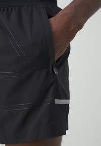 Mano derecha introducida en el bolsillo con cremallera de unos pantalones cortos deportivos negros con una franja reflectante y un patrón geométrico sutil.