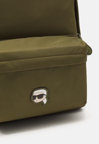 Borsa in nylon verde oliva con una texture liscia, dotata di un manico superiore cucito e di una toppa a forma di volto di cartone animato che indossa occhiali da sole sulla parte anteriore.