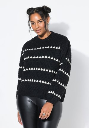 Frau mit Doppelknoten-Frisur, die einen schwarzen Strickpullover mit weißen Mustern und schwarze Lederimitat-Hosen vor einem einfarbigen Hintergrund trägt.