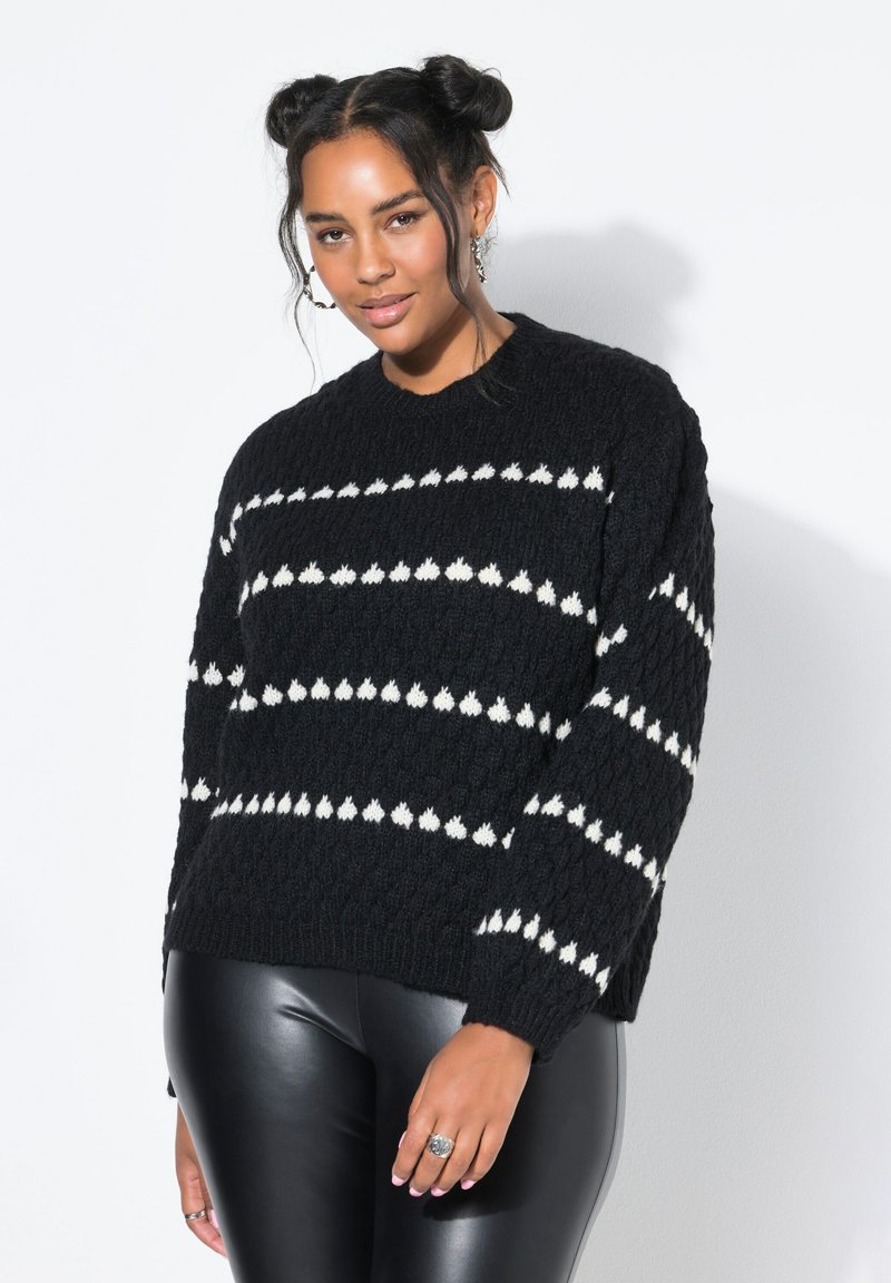 Donna con acconciatura a doppio chignon che indossa un maglione nero a maglia con motivi bianchi e pantaloni in ecopelle neri, su uno sfondo neutro.