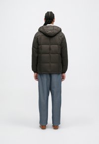 Mørkegrønn pufferjakke med hette, kviltet design og elastiske mansjetter, parret med lyseblå vide bukser og brune sko.