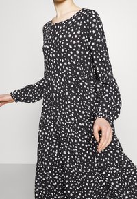 Musta mekko, jossa on valkoinen kukkakuvio, avonainen kaula-aukko, pitkät hihat ja kerroksellinen hameosa. Valmistettu pehmeästä kankaasta, virtaava muoto.