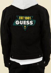 Svart denimjacka med en stor grön och gul broderad triangelpatch som står "EST 1981 GUESS?" på baksidan. Har knappar och en huva.