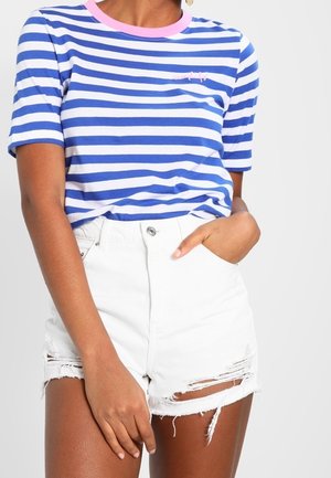 Kvinde iført blå og hvide stribede kortærmede skjorte stoppet ned i hvide højtaljede slidte denimshorts, med den ene hånd i lommen.