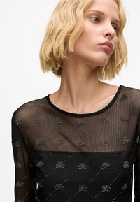 Schwarzes Mesh-Oberteil mit transparentem Design und silbernen Verzierungen, die in einem diagonalen Muster angeordnet sind. Runde Halslinie und lange Ärmel.