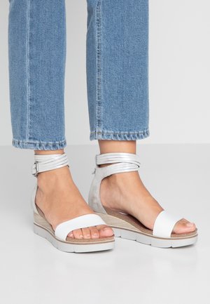 Fødder iført hvide kilehælssandaler med ankelremme, kombineret med lyseblå korte jeans mod en ensfarvet lys baggrund.