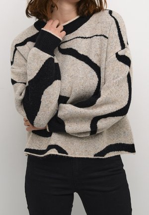 Femme portant un pull beige avec des motifs abstraits noirs et un pantalon noir, main touchant le cou, fond blanc.