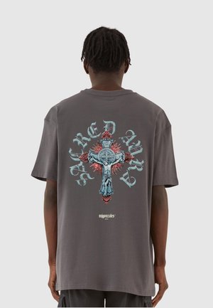 Grå bomulds t-shirt med et stort grafik af et kors med detaljerede illustrationer og farverige accenter på ryggen. Løs pasform og korte ærmer.